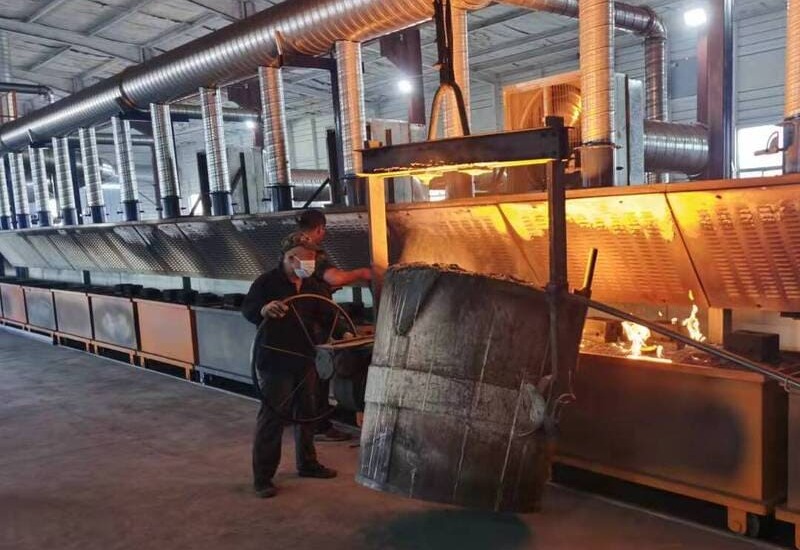 A man works on a large metal barrel in a metal casting foundry, surrounded by tools and equipment
